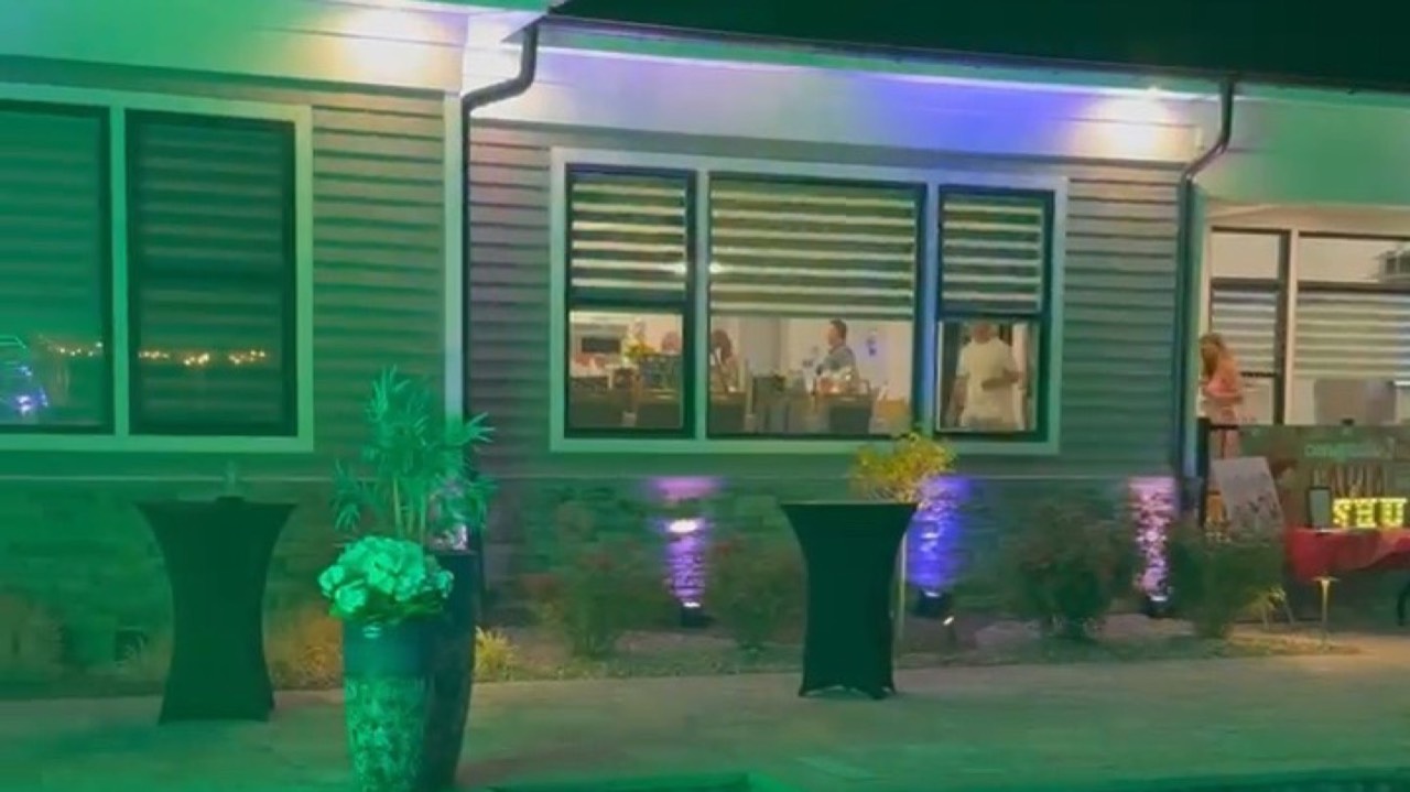 A back garden at night under purple uplighting on the house and a green wash across the pool deck, with cocktail tables set out