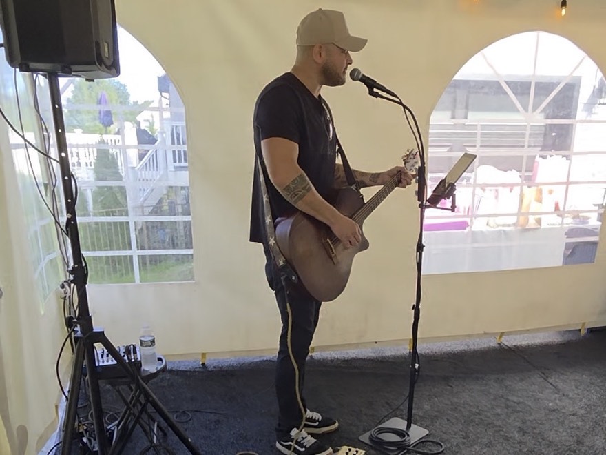 An acoustic guitarist singing into a microphone under a marquee, with a PA speaker and monitor set up beside him